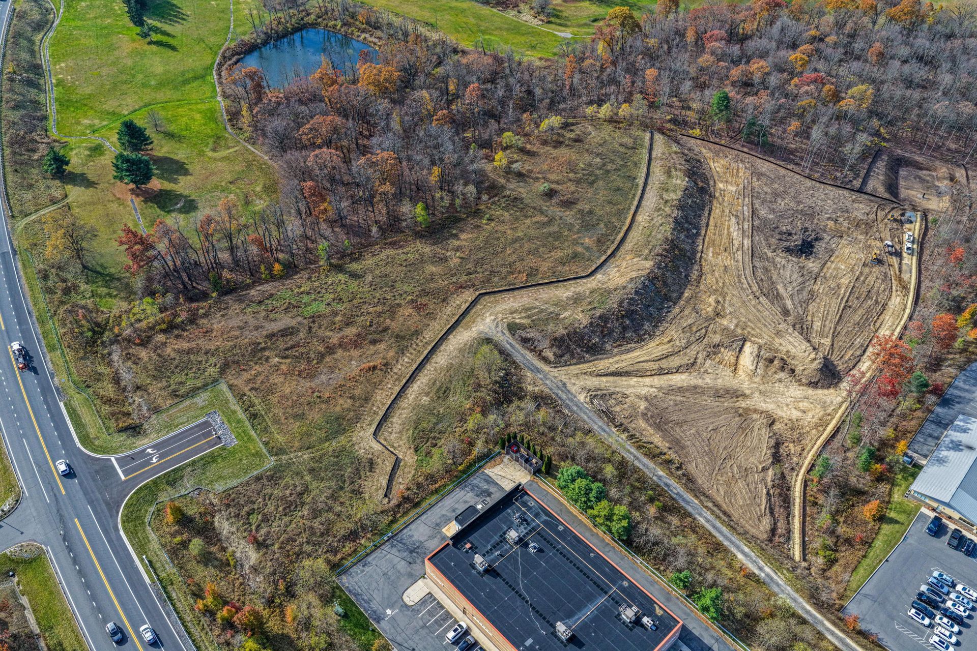 Aerial view of a construction site next to a road, field, and woods, with a pond visible.