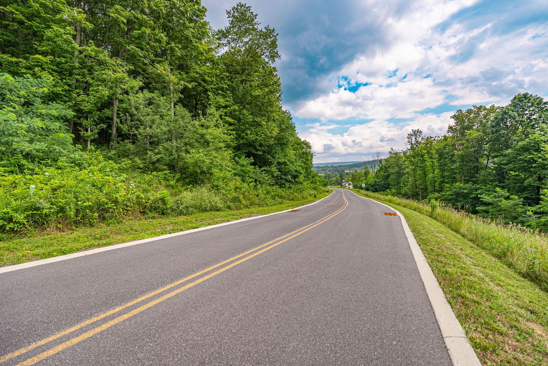 A paved road curves through a lush, green forest under a partly cloudy sky.