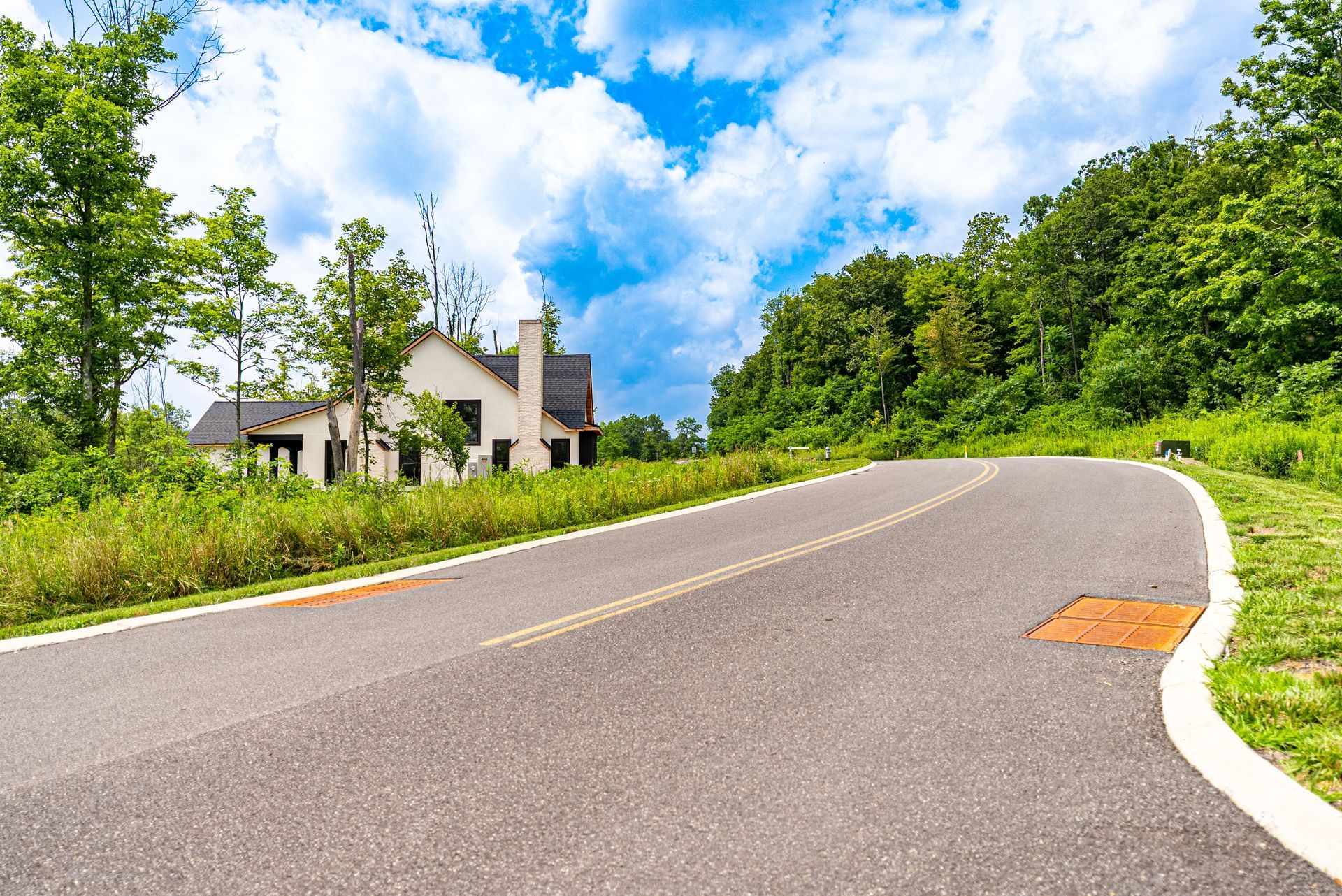 Paved road curves uphill toward a house under construction, bordered by grassy areas and a forested hillside.