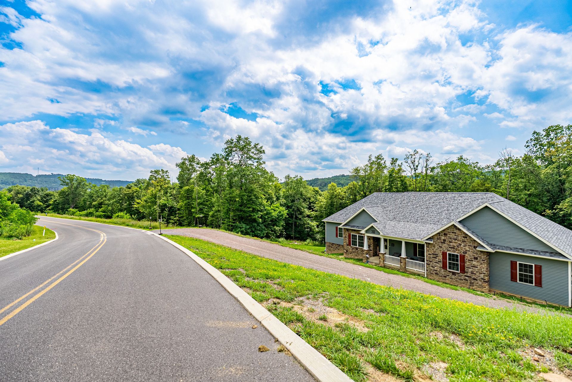 A house with a stone facade and gray siding sits beside a winding road. Green grass lines the road with trees.