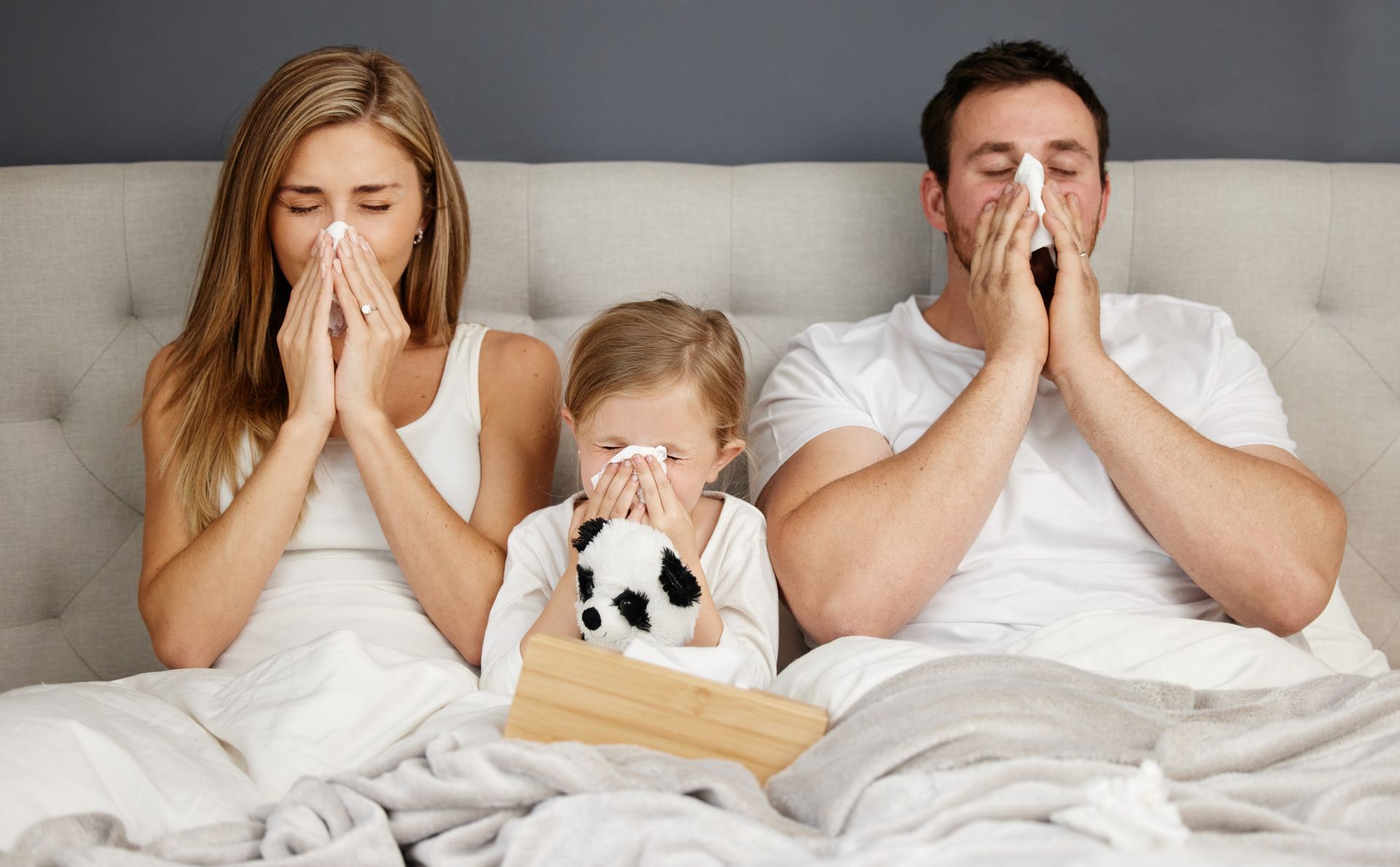 A family with sinus infection is sitting on a bed blowing their noses.