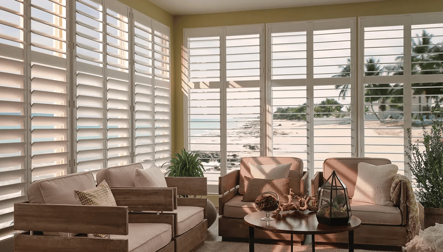 Bright living room with beige sofas, glass coffee table, and large windows with white blinds overlooking outdoors