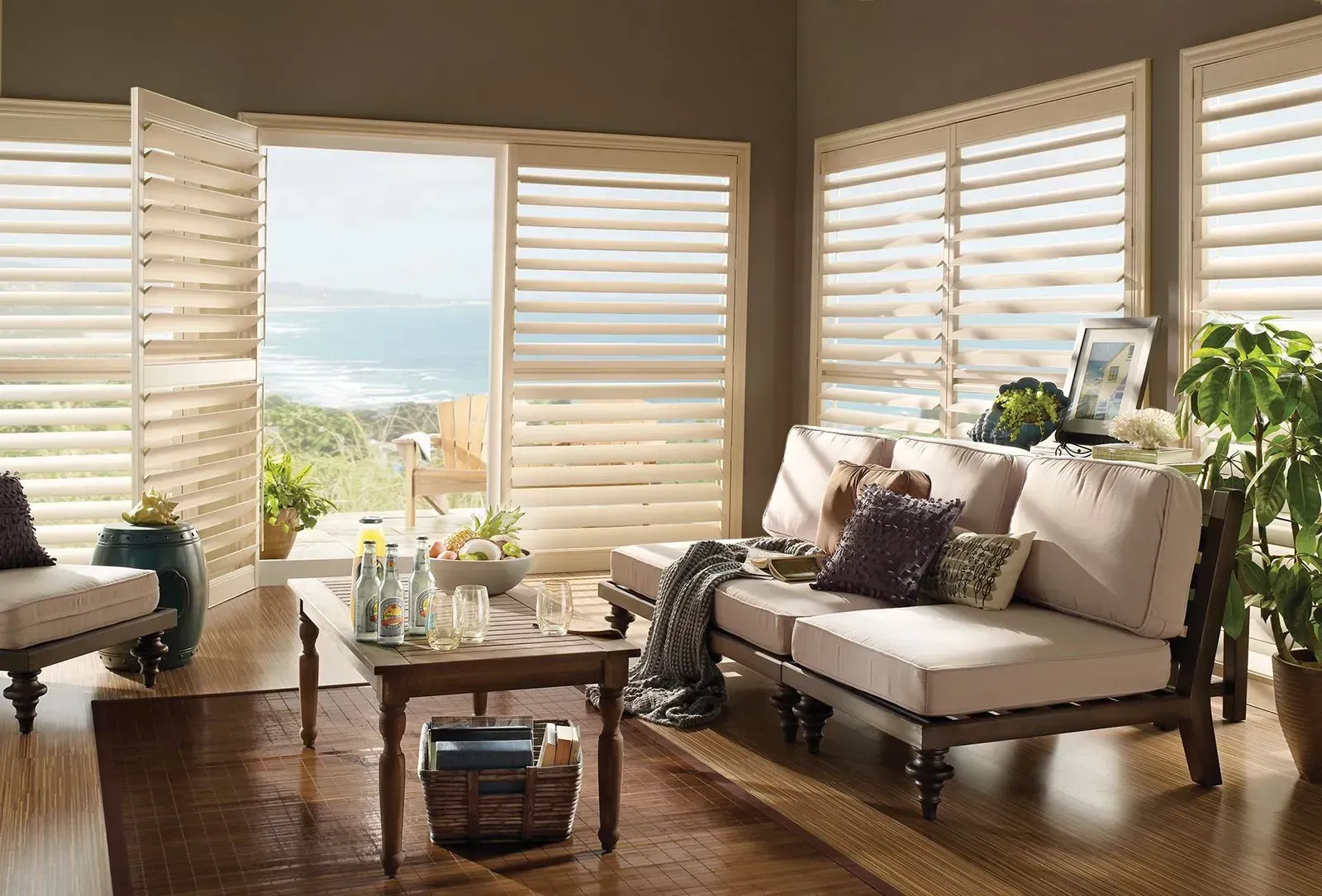 Bright living room with white shutters, beige sofa, wood coffee table, and ocean view through open windows