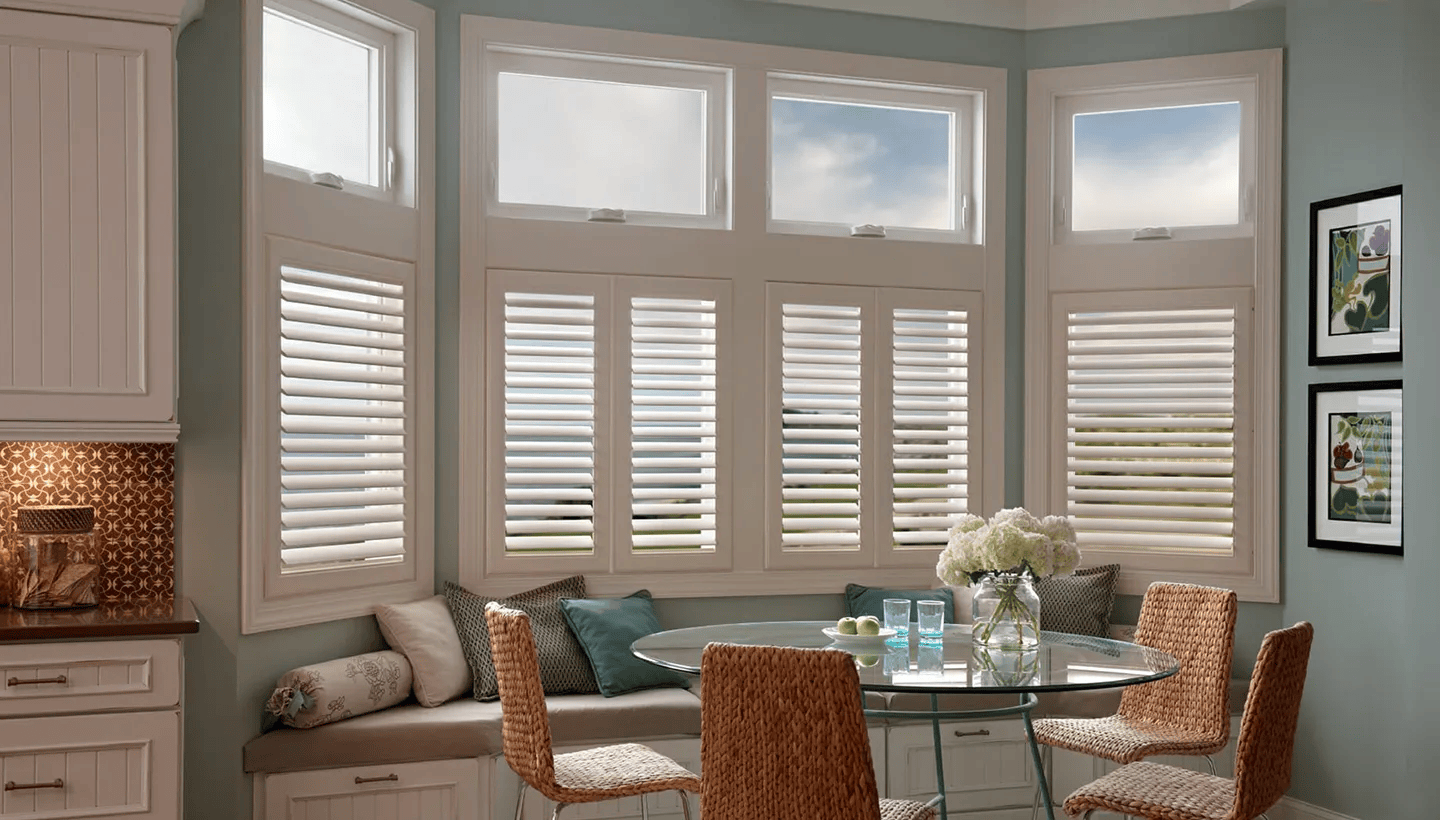 Bright breakfast nook with bay windows, white shutters, wicker chairs, and a glass table set with flowers.