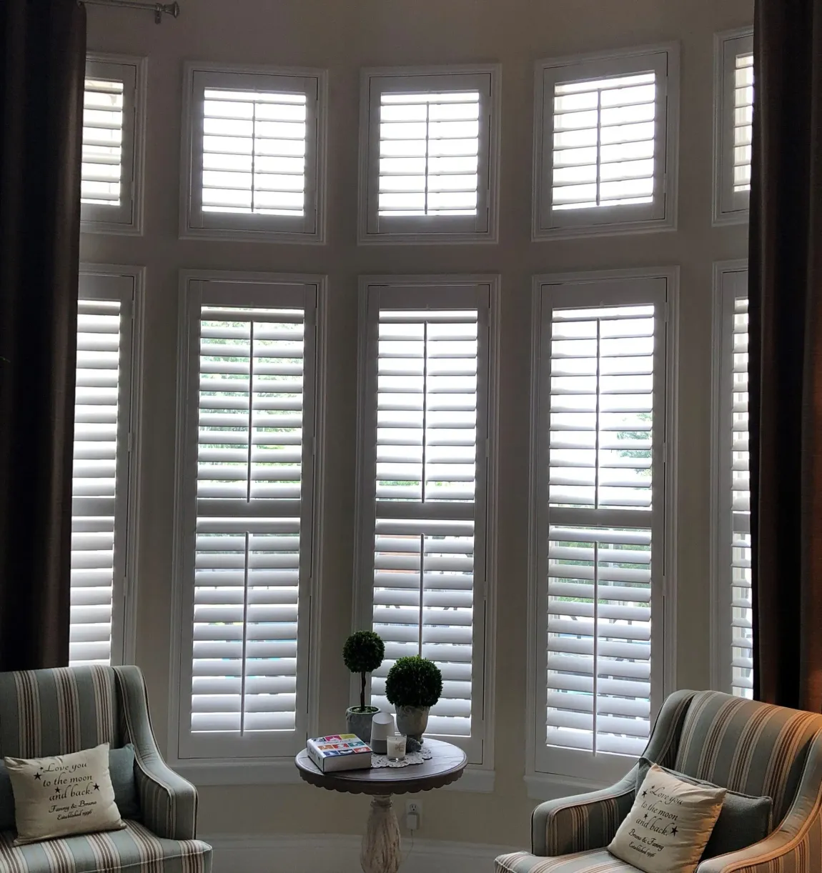 Sunlit bay window with white shutters, two striped armchairs, and a small round table with decor