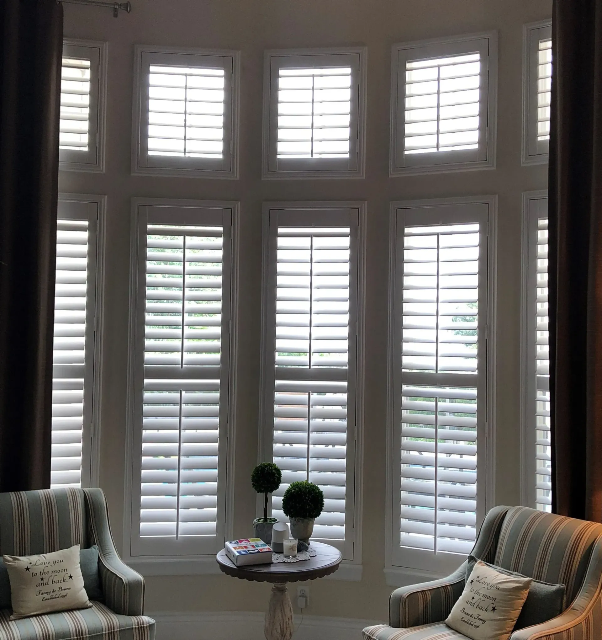 Sunlit bay window with white shutters, two striped armchairs, and a small round table with decor
