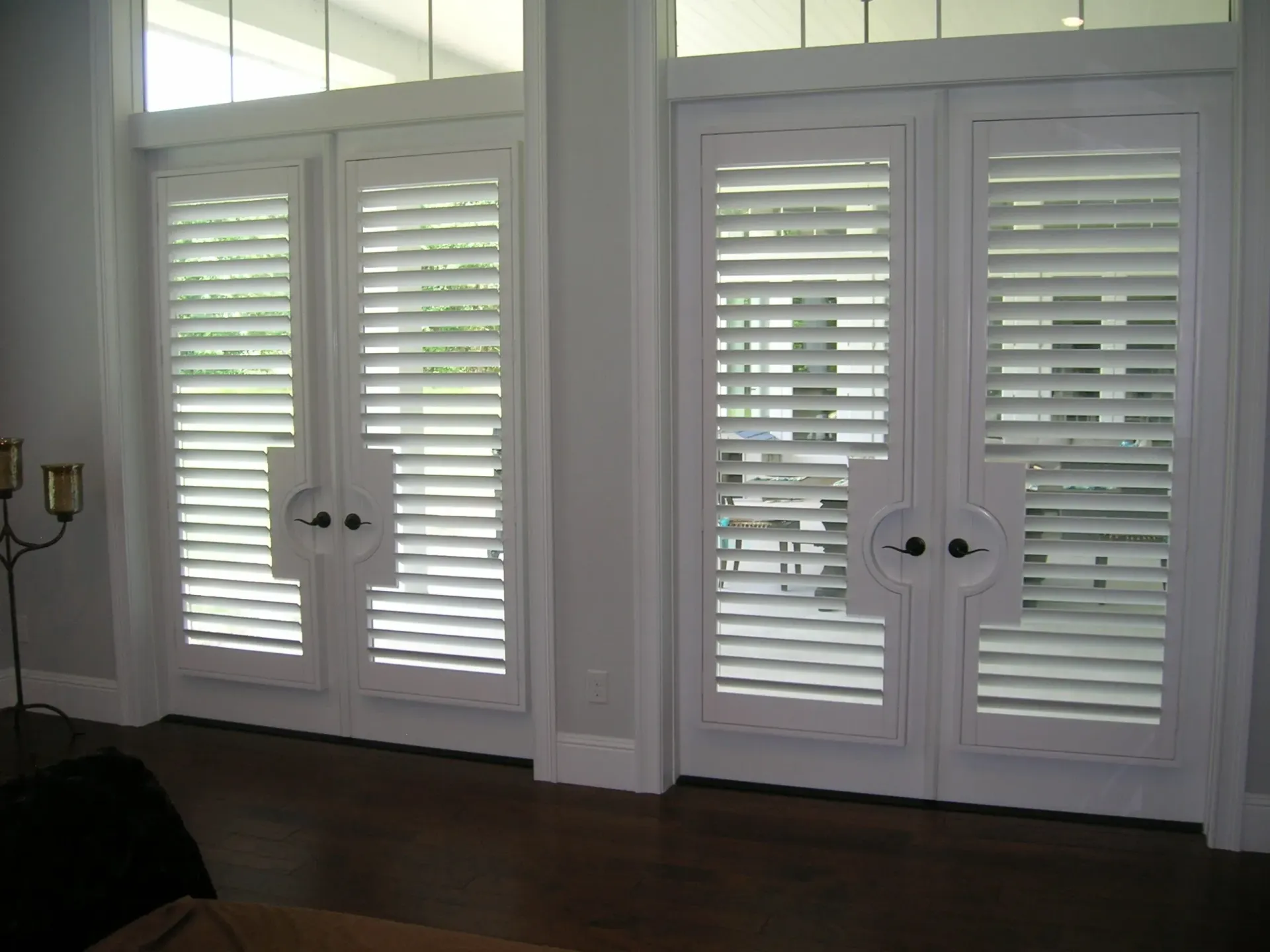 White plantation shutters on double doors in a bright room, with a dark floor and candle holder at left.
