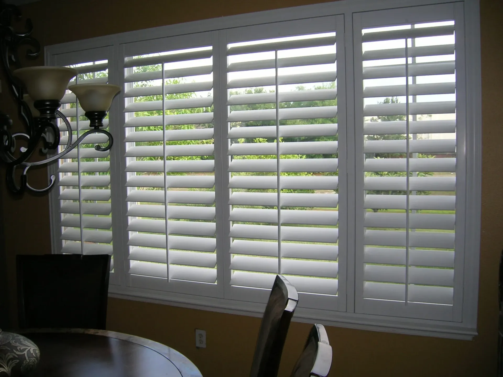 Dining room with white window shutters and a chandelier, showing greenery outside.