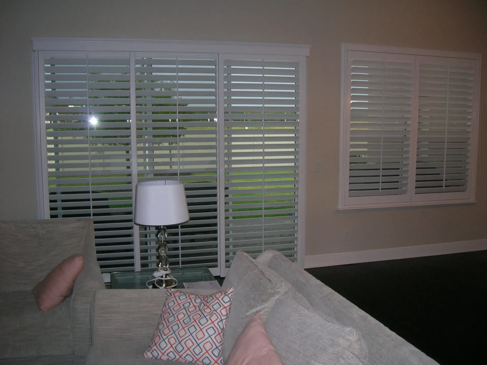 Living room with gray couch, white blinds, lamp, and a view of a green yard outside.