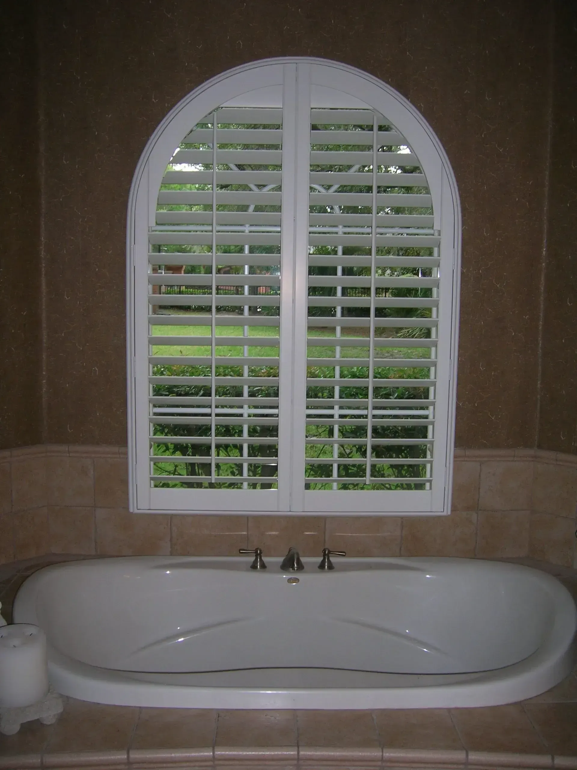 Bathroom with a large arched window, white shutters, and a freestanding tub below.