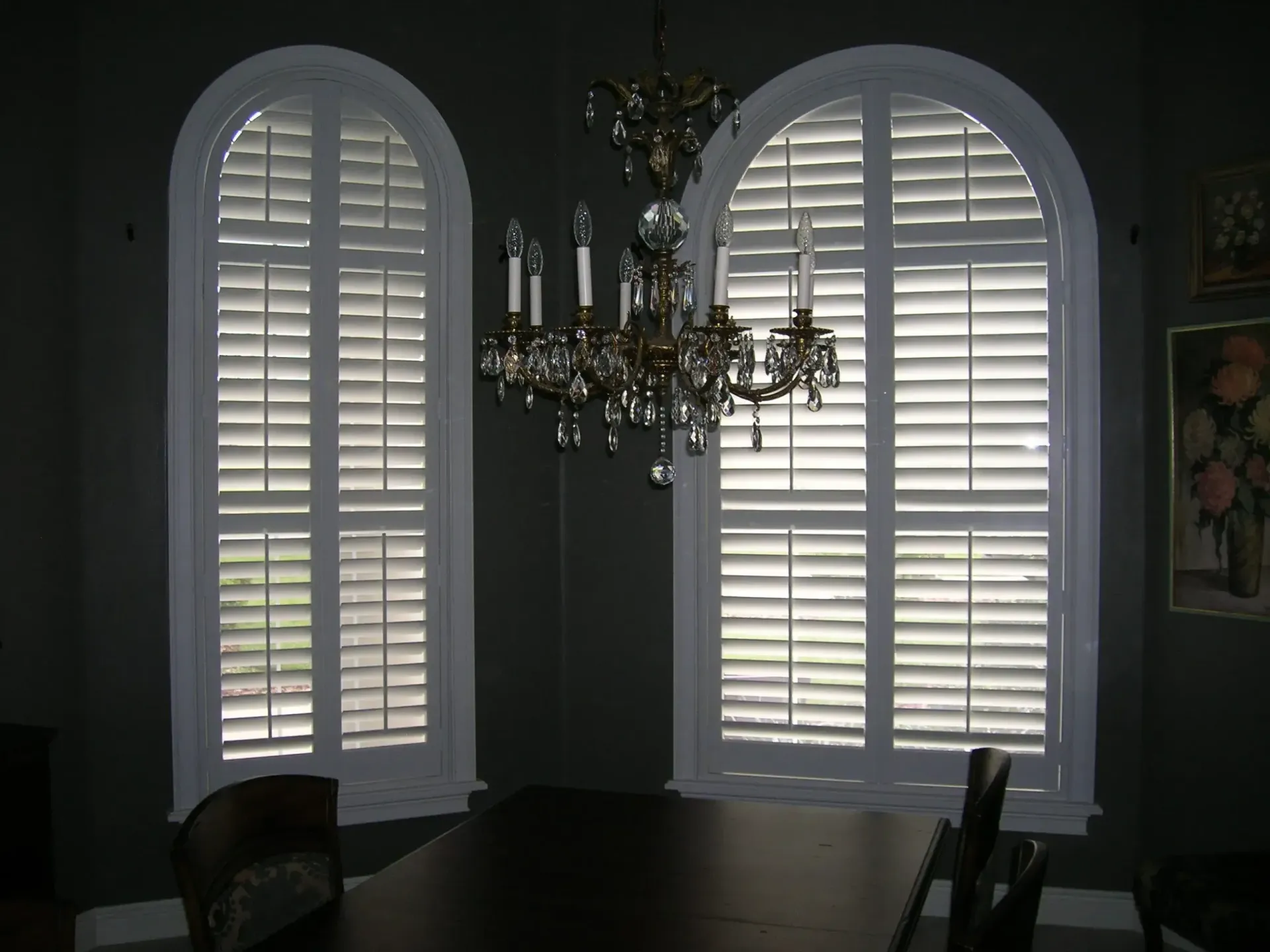 Dim dining room with two shuttered arched windows and a chandelier over a dark table
