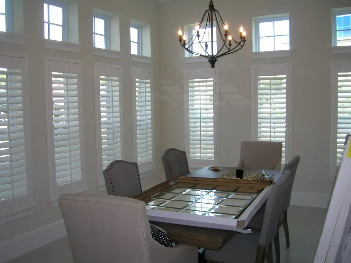Dining room with a glass-top table, upholstered chairs, tall white windows, and a hanging chandelier.