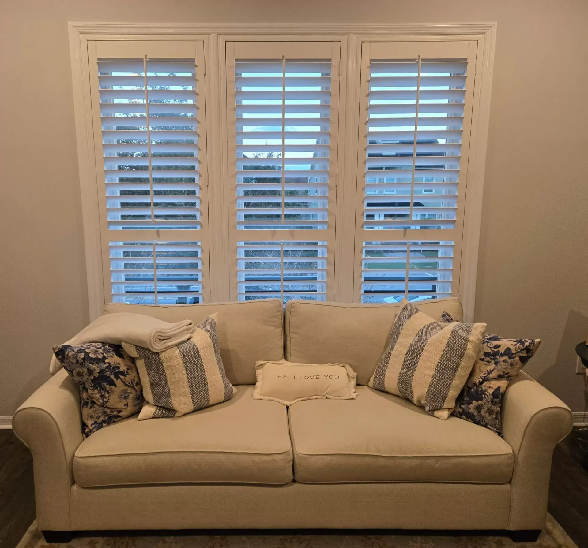 Cozy beige sofa with patterned pillows under white shutters in a softly lit living room.