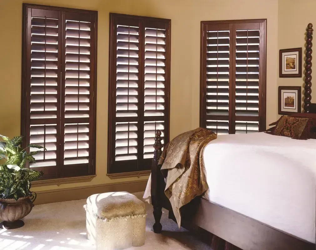 Bedroom with dark wood shutters, a bed, ottoman, potted plant, and warm beige decor.