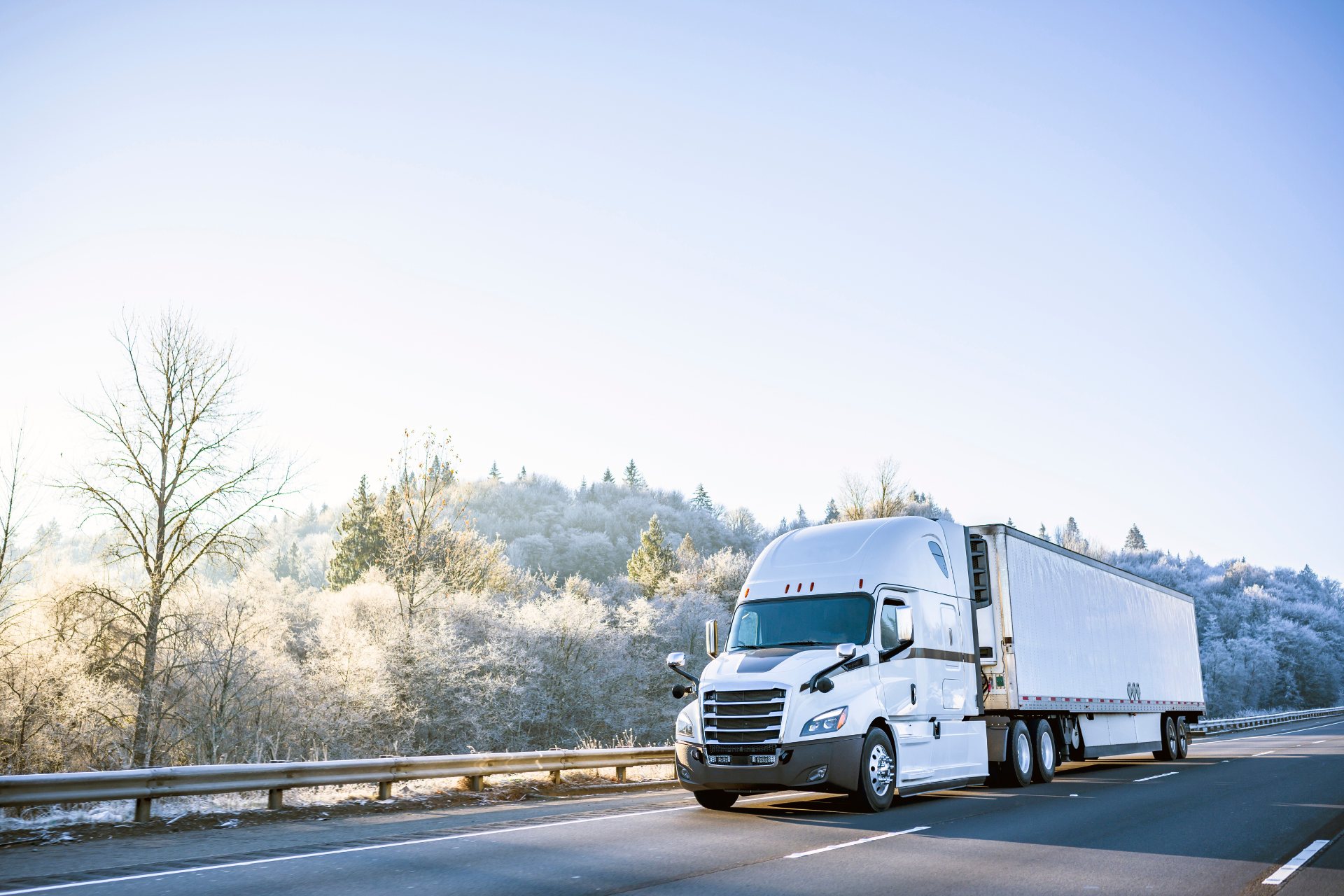 White refer semi-truck driving on a road with frosted trees and blue sky in the background.