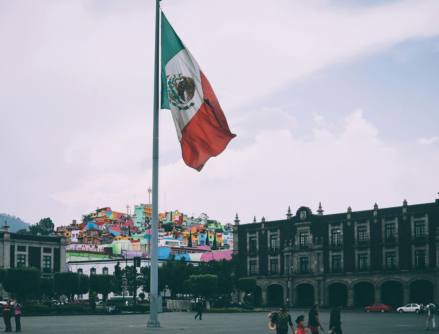Mexican flag waving in a square, colorful hillside houses, and a historic building.
