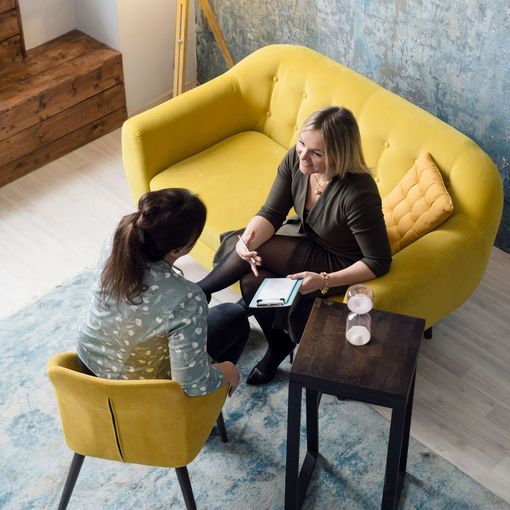 Two women are sitting on a yellow couch talking to each other.