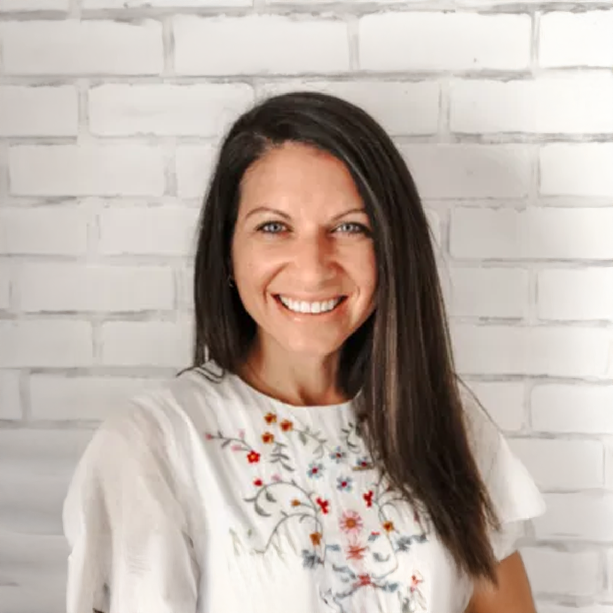 A woman in a white shirt is smiling in front of a white brick wall. A woman in a white shirt is smiling in front of a white brick wall.