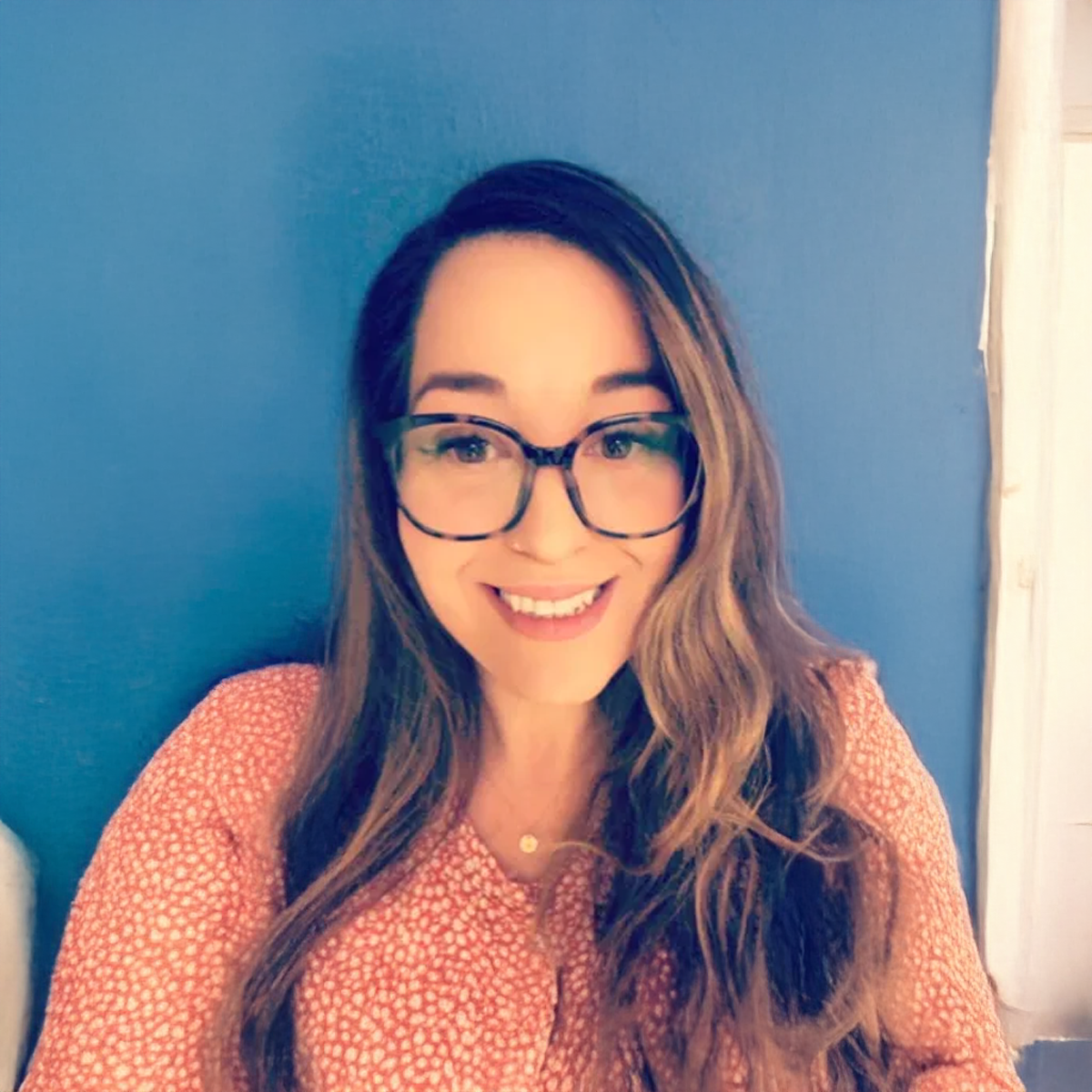 A woman wearing glasses and a pink shirt is smiling in front of a blue wall. A woman wearing glasses and a pink shirt is smiling in front of a blue wall.