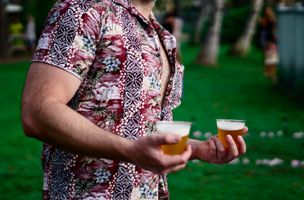 A man in a floral shirt is holding two cups of beer in his hands. A man in a floral shirt is holding two cups of beer in his hands.