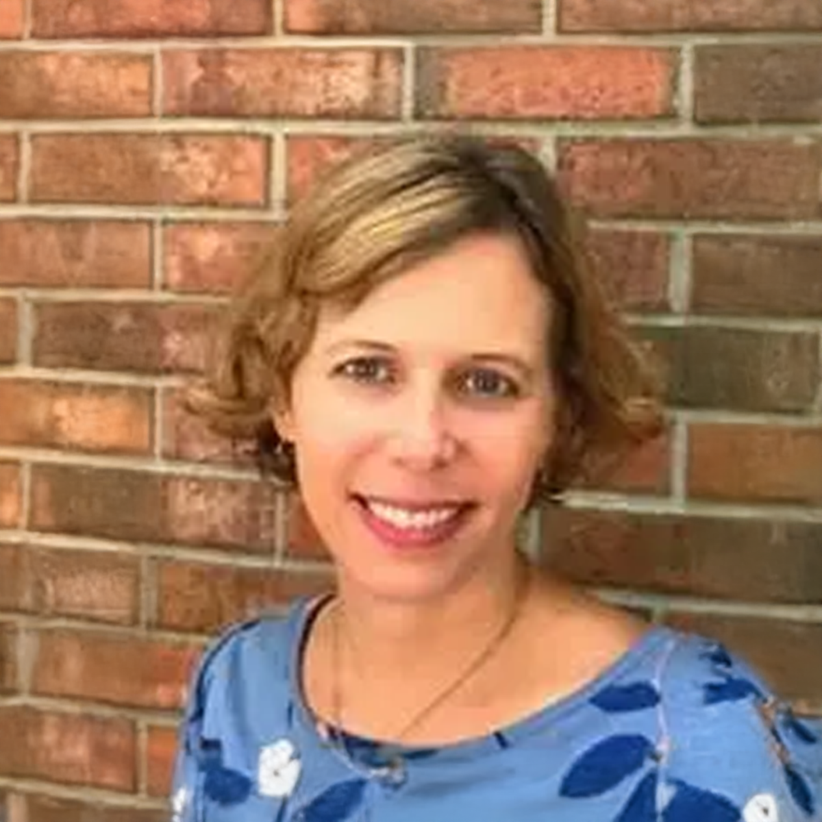 A woman in a blue shirt is smiling in front of a brick wall. A woman in a blue shirt is smiling in front of a brick wall.