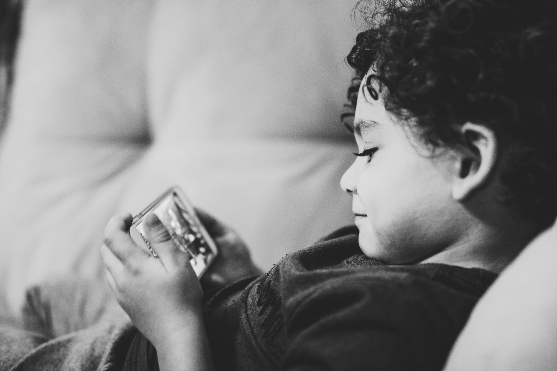 A young boy is laying on a couch playing with a cell phone. A young boy is laying on a couch playing with a cell phone.