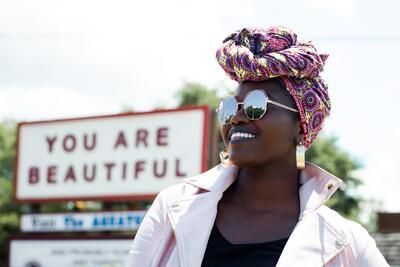 A woman wearing sunglasses and a head scarf is standing in front of a sign that says `` you are beautiful ''.