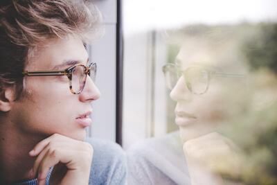 A young man wearing glasses is looking out of a window.