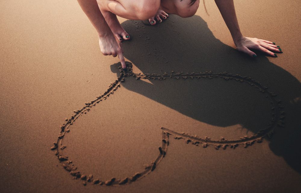 A woman is kneeling on the beach and drawing a heart in the sand. A woman is kneeling on the beach and drawing a heart in the sand.