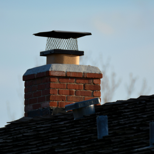 Brick chimney with metal cap on a dark roof against a light blue sky.