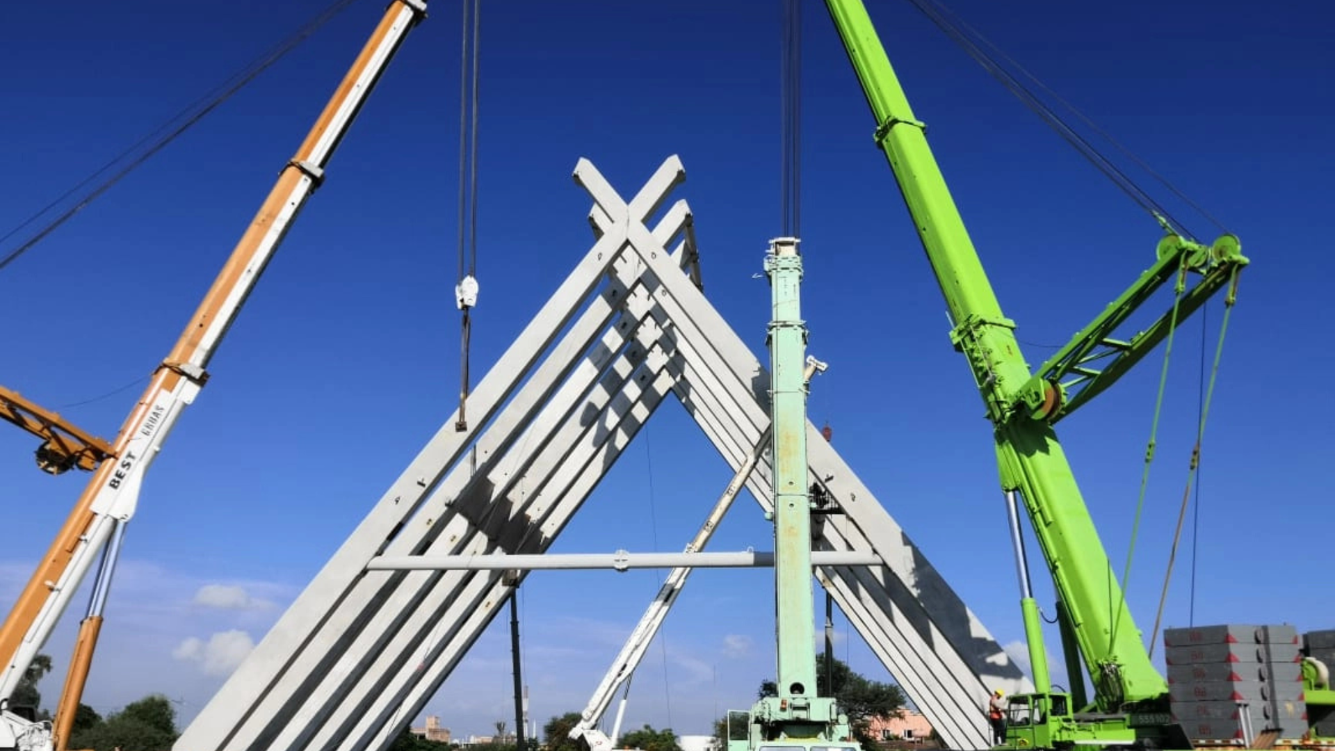 Cranes lifting and assembling a white triangular structure against a blue sky.