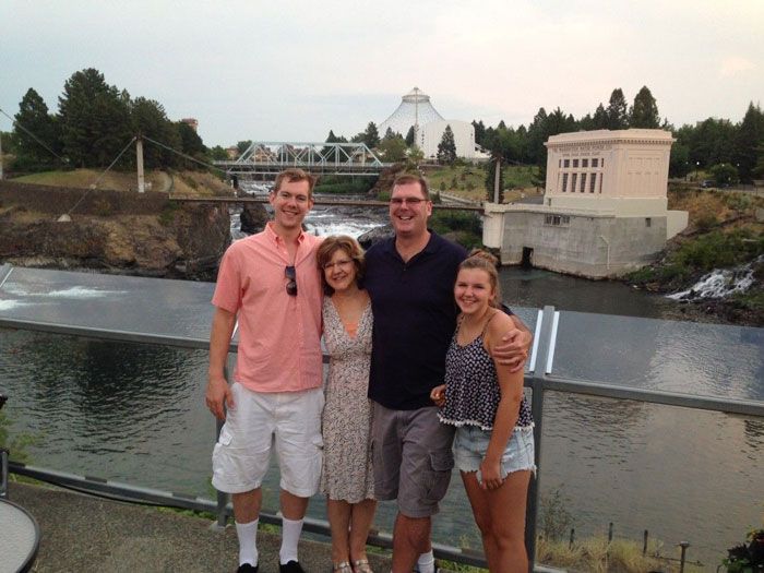 Four people pose near a river with a dam and a bridge in the background.