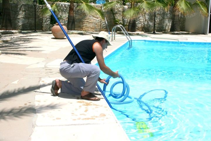 Person kneeling by a pool, using a blue hose to clean the water