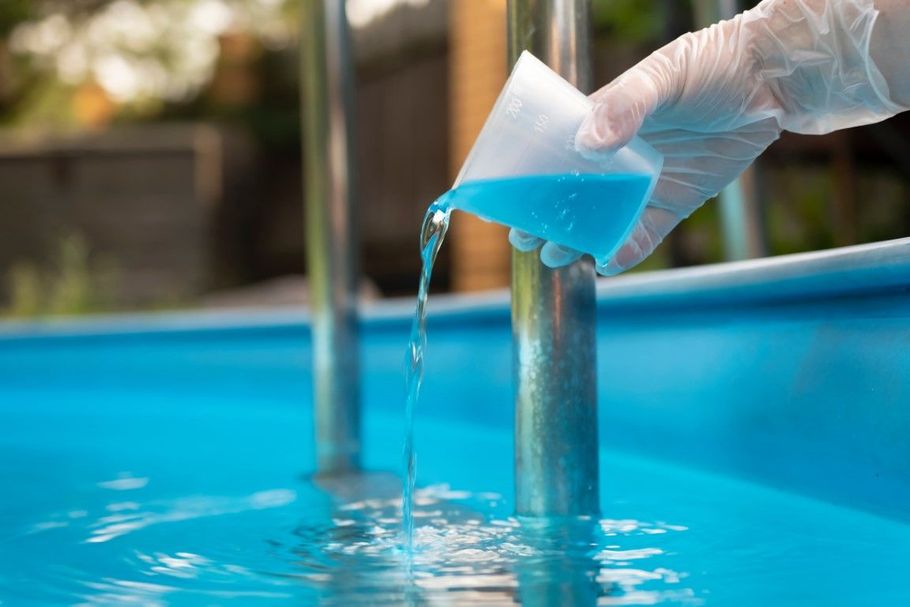 Gloved hand pouring blue pool test liquid into a water sample by a swimming pool