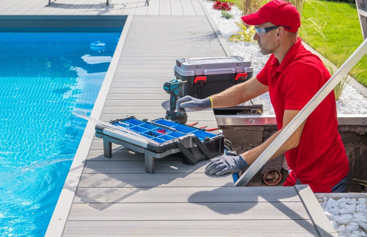 Pool worker in red uniform cleaning the poolside deck beside turquoise water.