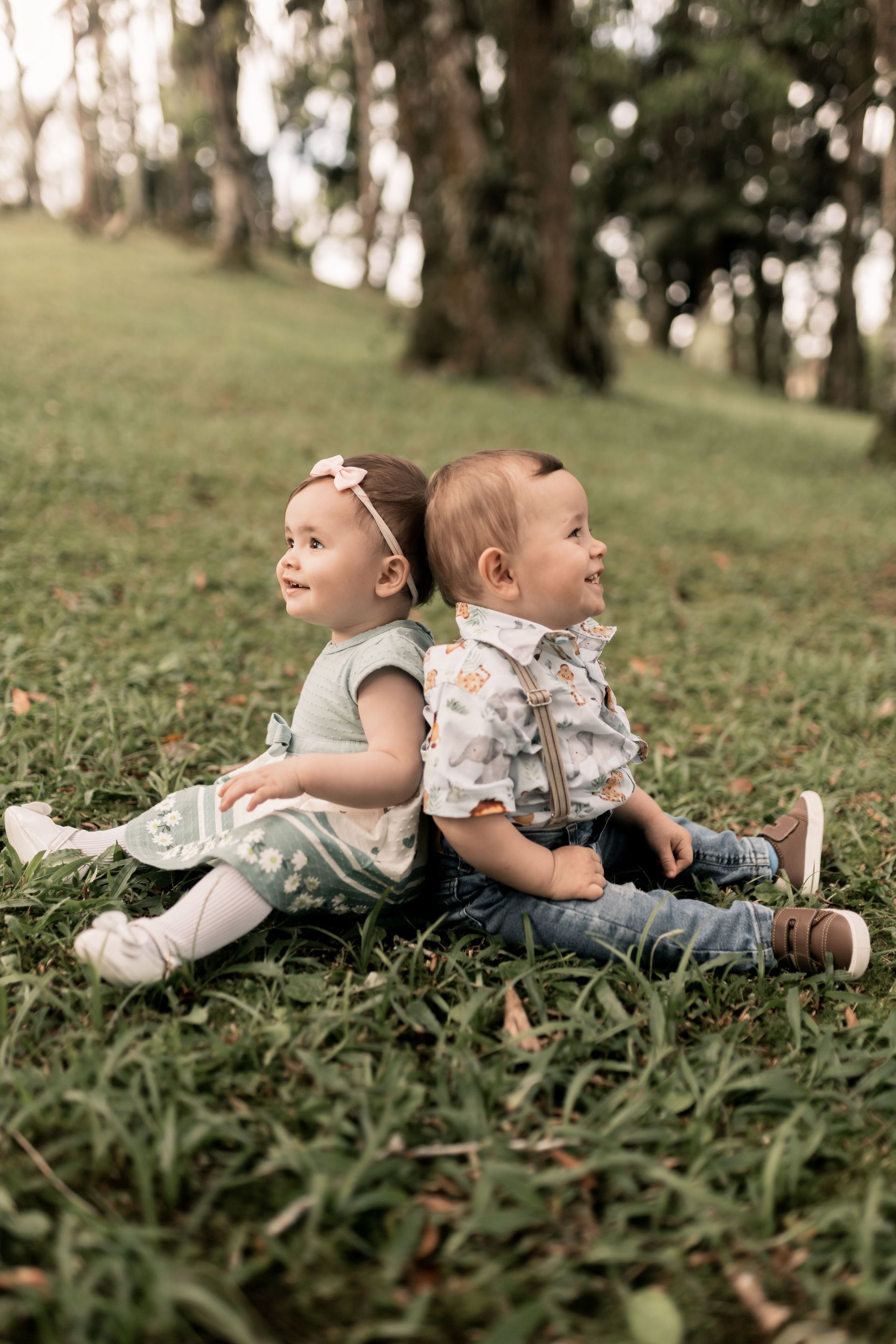 Due bambini piccoli seduti schiena contro schiena sull'erba di un parco, che guardano in direzioni opposte, con gli alberi sfocati sullo sfondo.