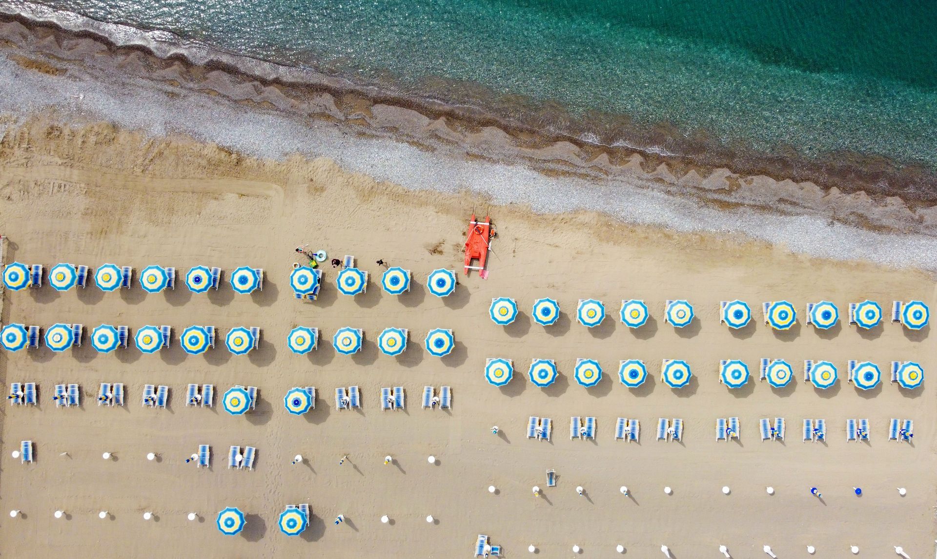 Veduta aerea di una spiaggia con file di ombrelloni e sedie blu sulla sabbia accanto a un'acqua turchese.