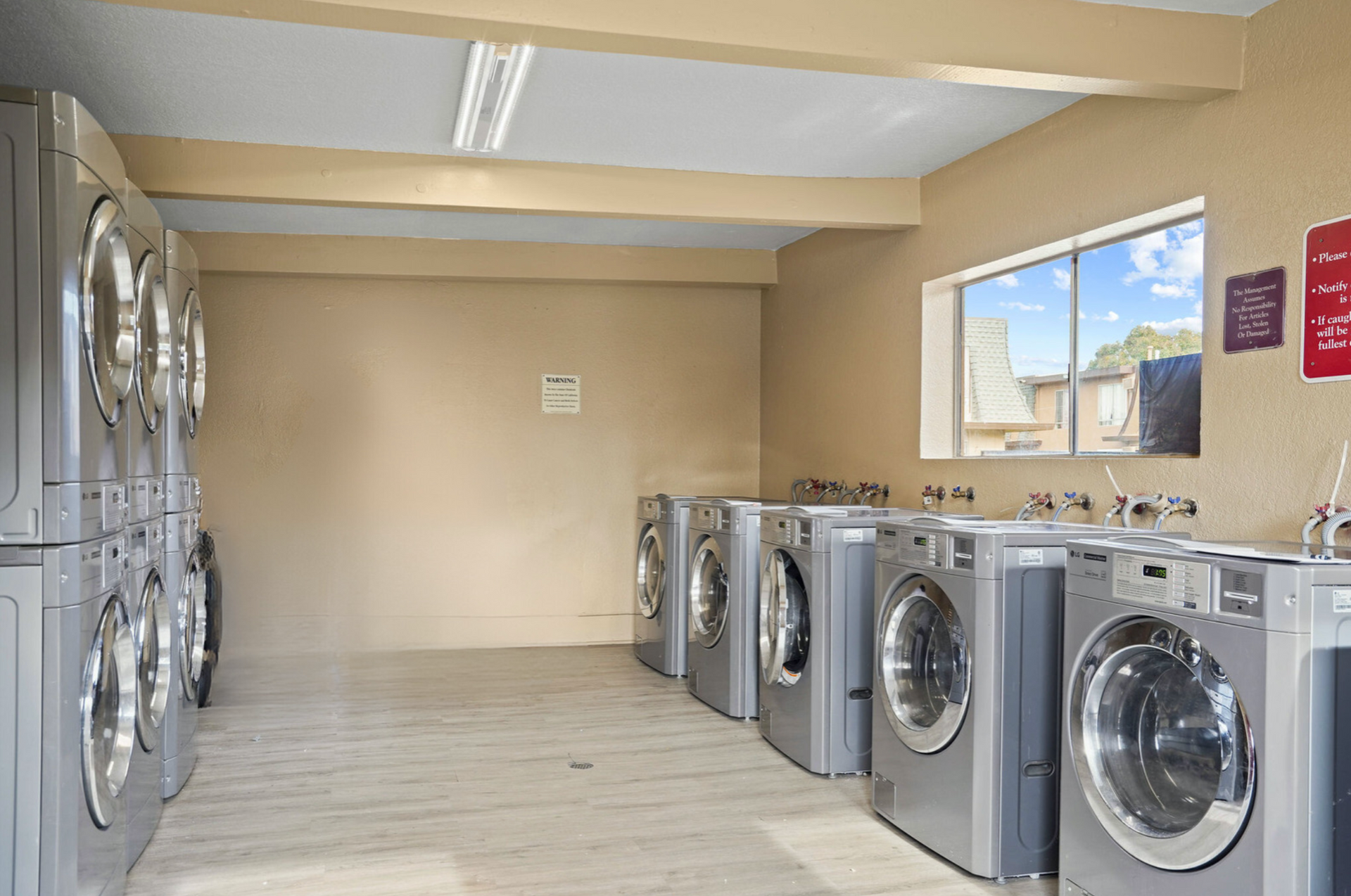 Laundry room with rows of silver washing machines and dryers along beige walls and a window.