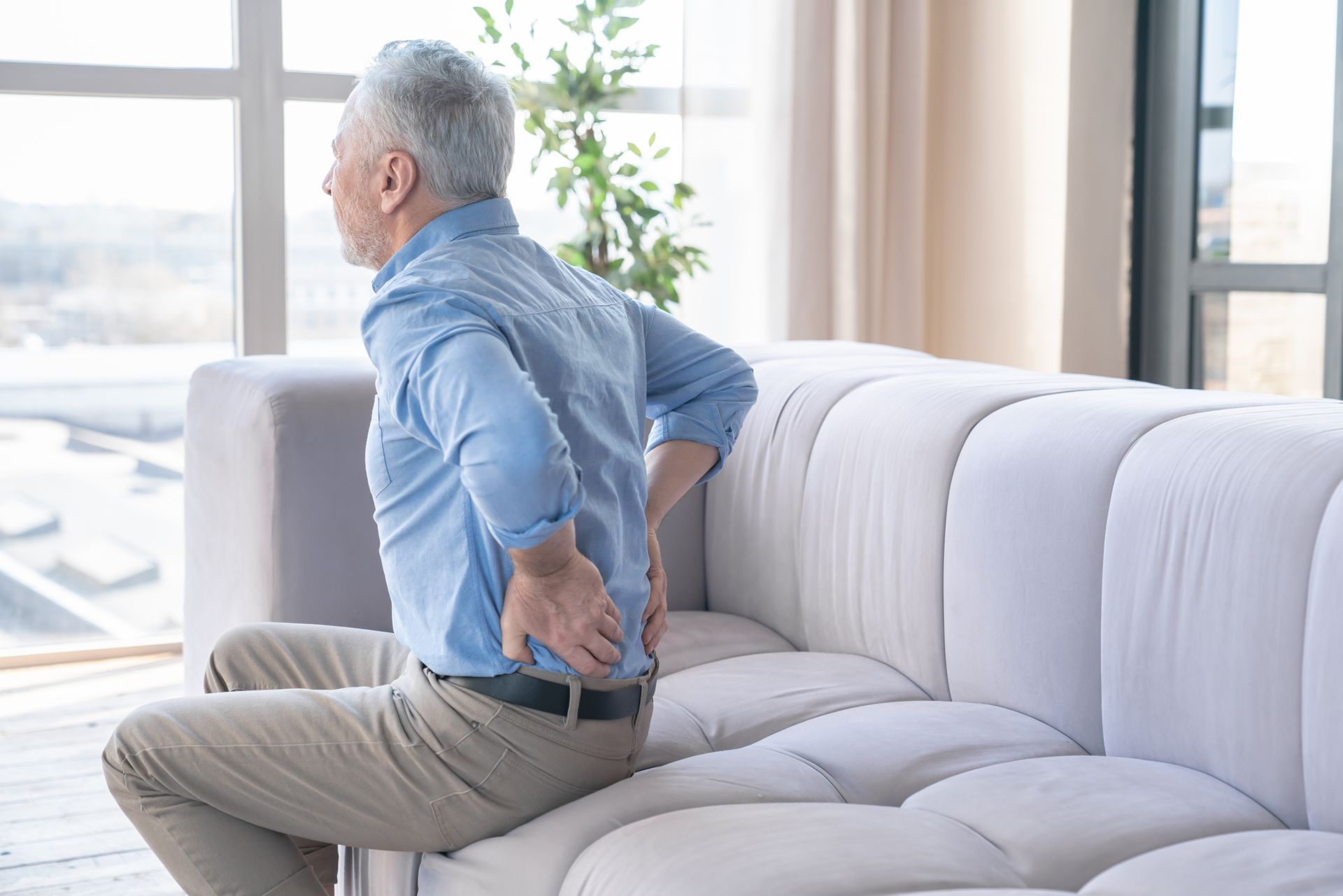 Man holding his lower back while sitting on a couch in a bright living room