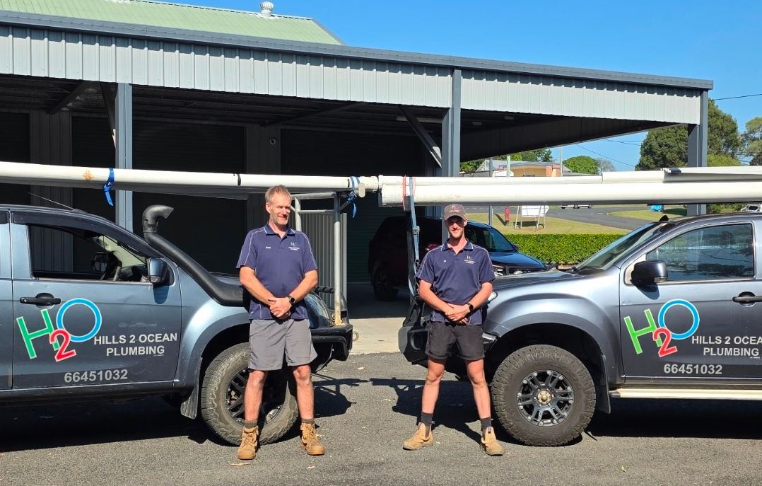 Two workers standing between two grey utility trucks in front of a shed, with tools and signage on the doors Hills 2 Ocean Plumbing in Maclean, NSW