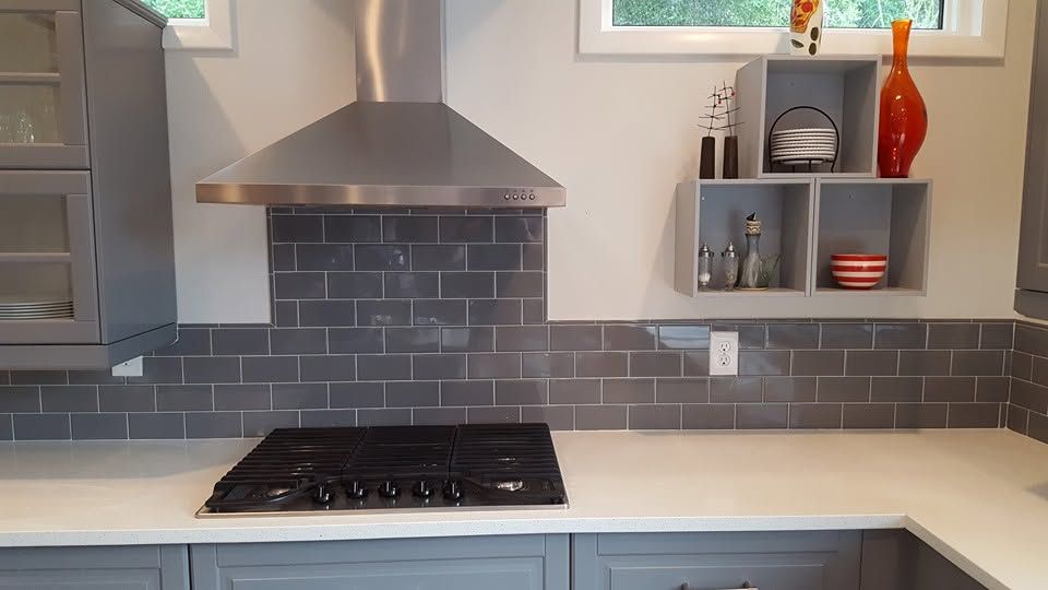 Modern kitchen with gray tile backsplash, stainless range hood, white counters, and open shelving.