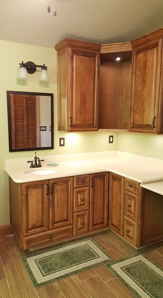 Wooden bathroom vanity with white countertop, mirror, wall sconce, and two green rugs in a corner sink area