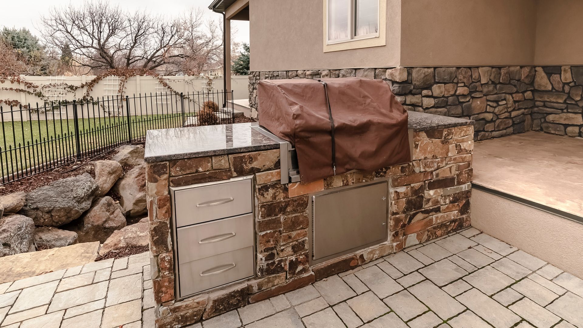 Stone mailbox and utility boxes beside a house with a covered grill, set on a paved driveway.