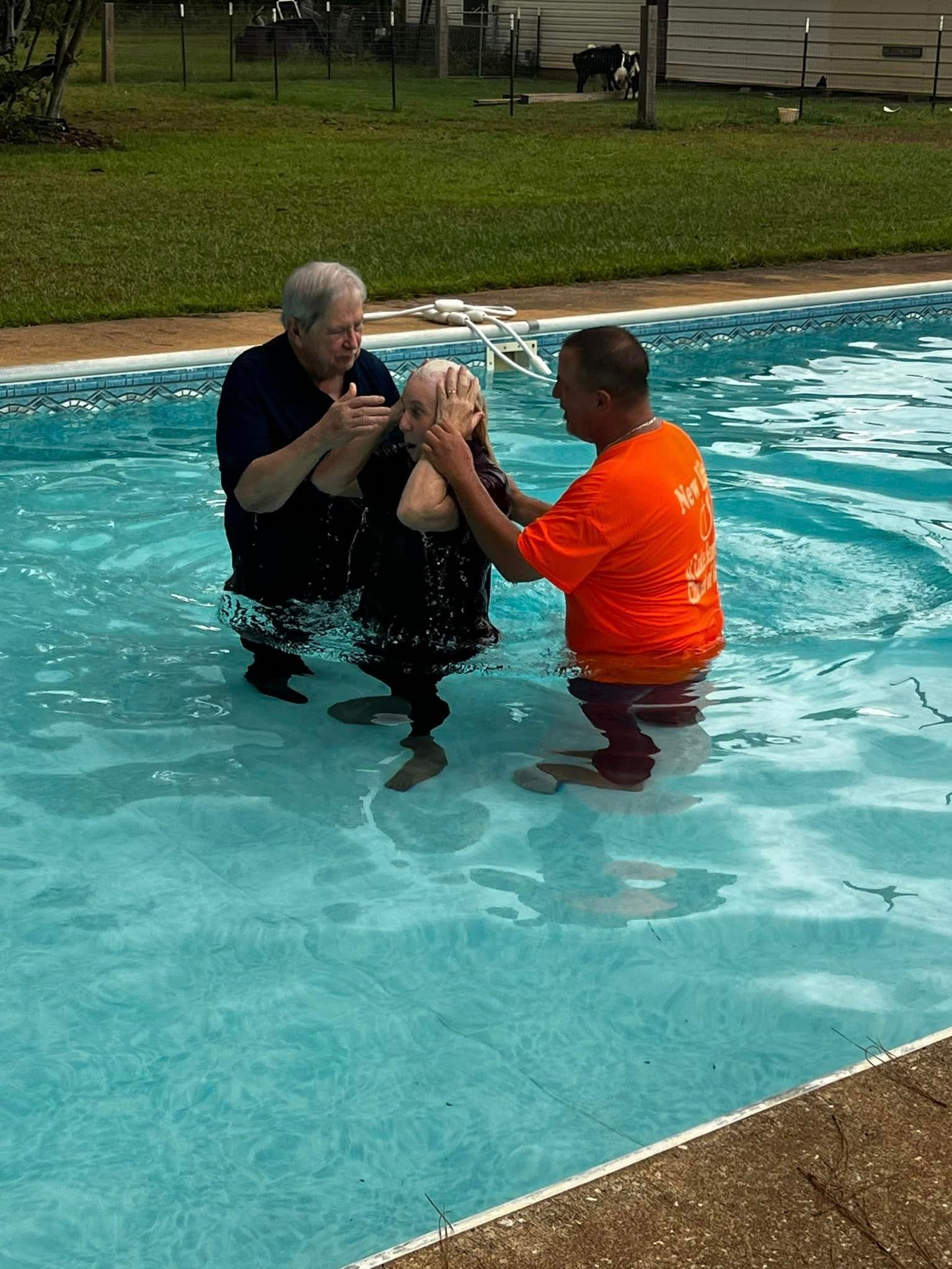 A man in an orange shirt is baptizing another man in a swimming pool.