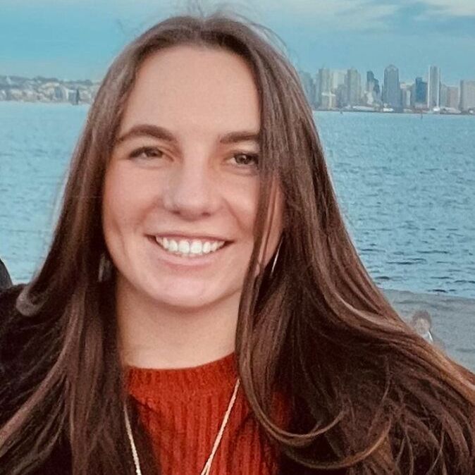A woman with long brown hair is smiling in front of a body of water.