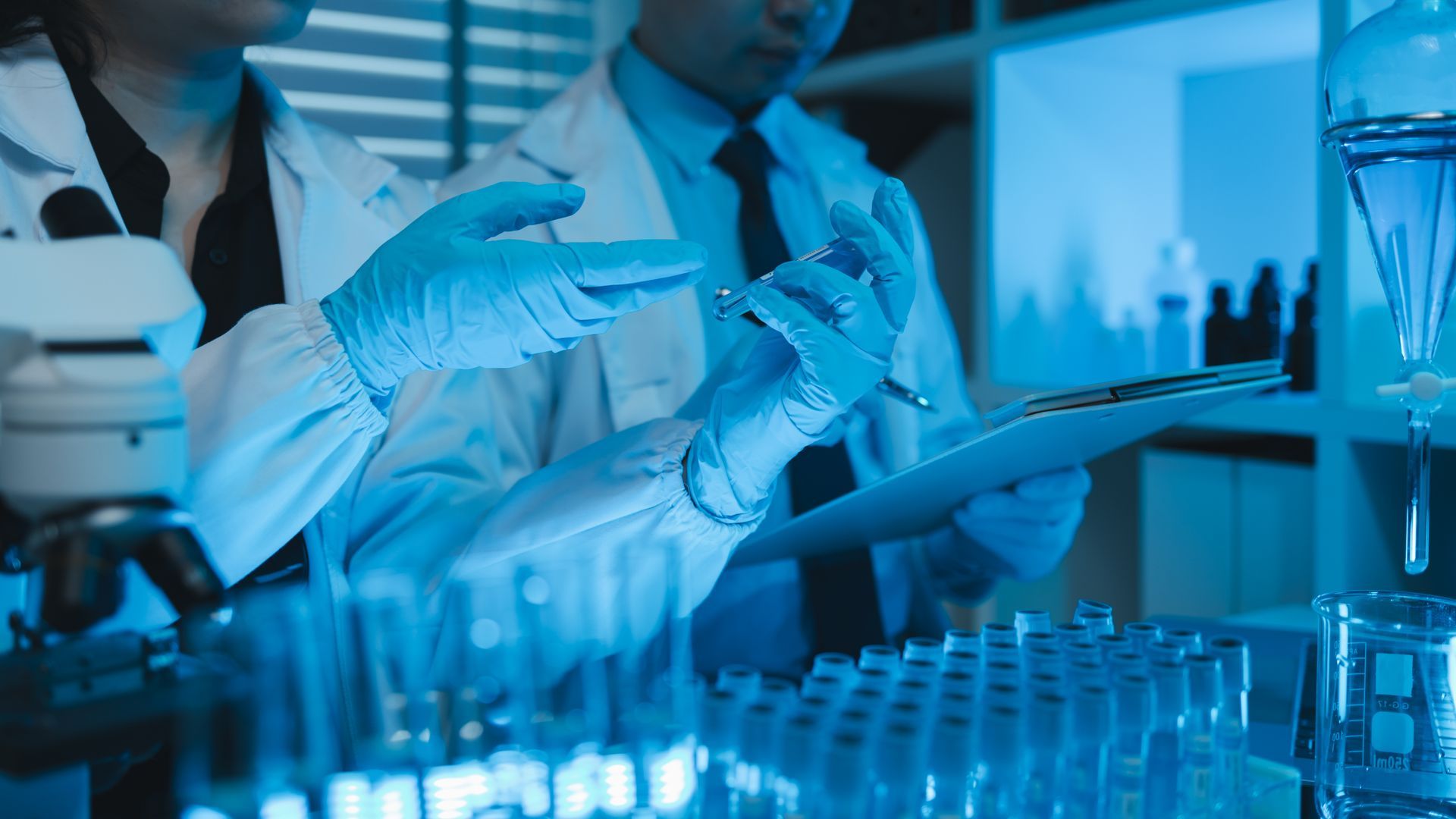 A man and a woman are working in a lab with a microscope and a clipboard.