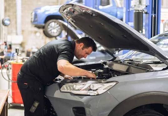 Mecánico inspeccionando el motor de un coche gris en un taller mecánico con el capó abierto.