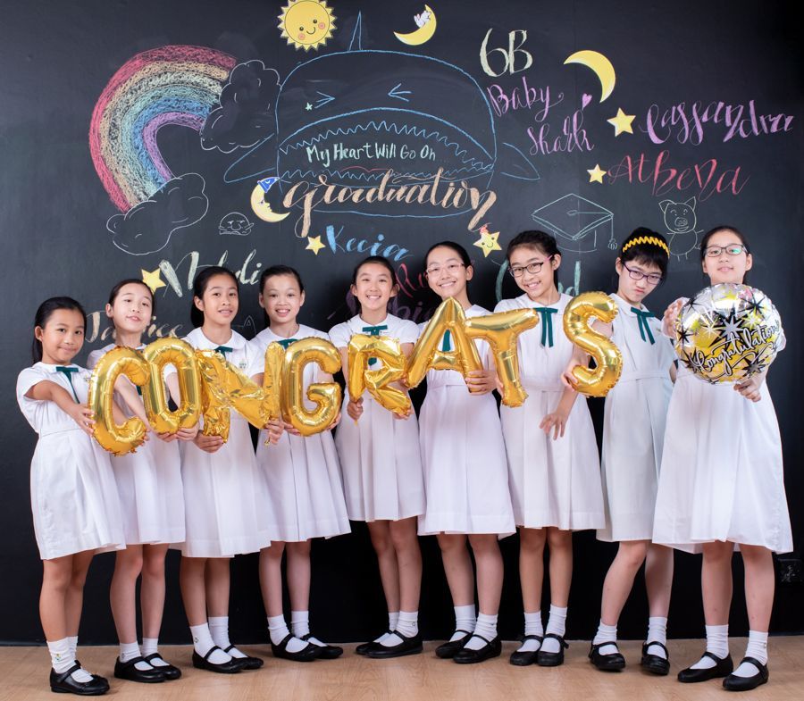 一群身穿白色洋裝的女孩手持金色「CONGRATS」氣球，在黑板前微笑。