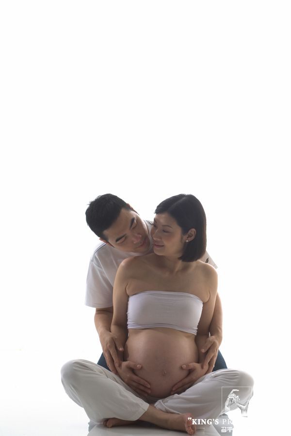 孕婦和伴侶，坐著，擁抱在一起。他用鼻子蹭著她的脖子，雙手放在她的腹部。白色背景，柔和的燈光。