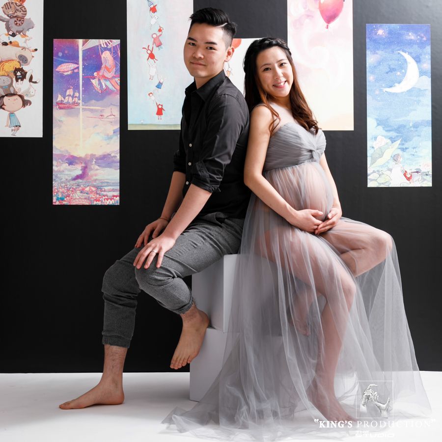 懷孕的夫婦背靠背坐著；男人穿著黑色襯衫，女人穿著灰色連身裙，靠著掛著藝術品的黑色牆壁。
